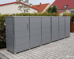 . Mehrere graue Paketboxen auf gepflastertem Boden, mit Hecken und Häusern im Hintergrund.