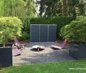 . Gartenbereich mit Liegestühlen, Feuerstelle und Pflanzen, umgeben von Bäumen und Hecken.