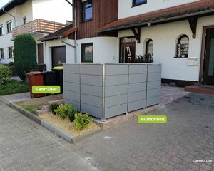 Graue Mülltonnenbox und Fahrradabstellplatz vor einem Haus mit gelber und roter Mülltonne.
