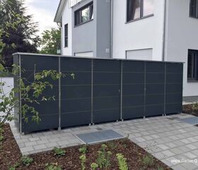 Großer grauer Gartenschrank neben einem modernen Gebäude, umgeben von Pflanzen und gepflastertem Boden.