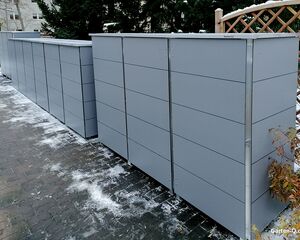 Graue Mülltonnenboxen auf einem gepflasterten Weg, umgeben von Schnee und Pflanzen.