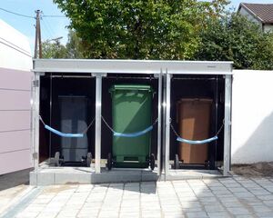 . Drei Mülltonnen in einem Metallgehäuse auf einer gepflasterten Fläche, jeweils mit blauen Gurten gesichert.