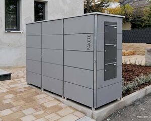 . Grauer Paketkasten mit mehreren Fächern steht draußen auf gepflastertem Boden.