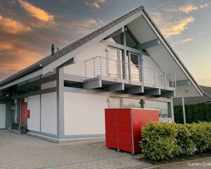 Ein modernes Haus mit Balkon, rotem Schrank draußen, Pflanzen und einem bedeckten Himmel im Hintergrund.