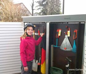 . Ein Kind mit Helm steht neben einem Gartenschrank voller Werkzeuge, darunter Harke, Schaufel, Rechen und eine Gießkanne.