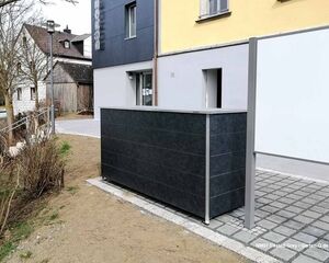 Ein moderner Mülleimerbehälter vor einem Gebäude mit gelber Fassade und umliegenden Häusern.