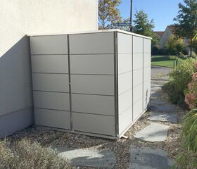 . Ein moderner, grauer Gartenschuppen steht an einem Gebäude, umgeben von Pflanzen und Kiesweg.