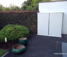 . Ein Gartenbereich mit Hecke, Busch, zwei grünen Pflanzentöpfen und einem weißen Gartenschrank an der Wand.