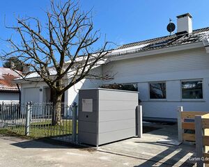 Ein modernes Haus mit kahlem Baum und grauem Zaun bei blauem Himmel.