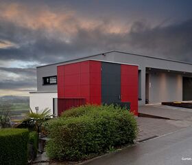Modernes Haus mit roter und grauer Fassade, umgeben von Büschen und dramatischem Himmel.