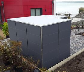 . Ein grauer Gartenschrank neben einem modernen Haus mit roter Fassade, umgeben von Pflanzen und Pflastersteinen.