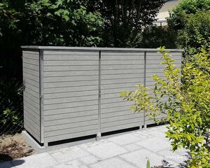 . Ein grauer Gartenschrank aus Holzimitat steht auf einer gepflasterten Fläche, umgeben von Büschen und Bäumen.