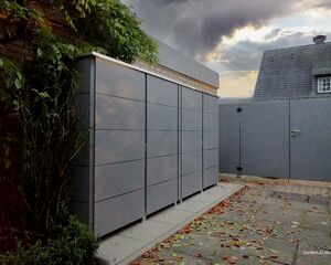 Ein modern gestalteter Gartenschrank neben einem Zaun, umgeben von Laub und Pflanzen.