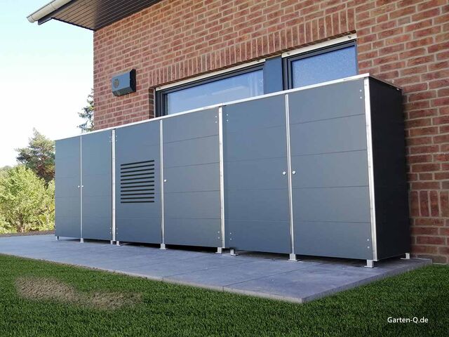 . Großer, grauer Metallschrank an der Hauswand, auf einer Terrasse mit Garten im Hintergrund.