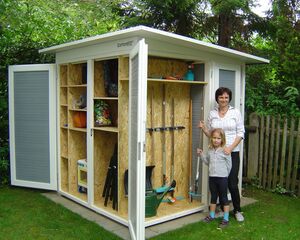 Eine Frau und ein Kind stehen vor einem geöffneten Gartenschuppen mit Gartenwerkzeugen und Spielsachen.