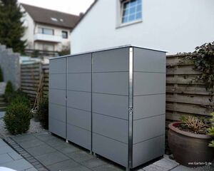 Ein graues Gartenschrank neben einem Holzzaun, umgeben von Pflanzen und einem Wohnhaus im Hintergrund.