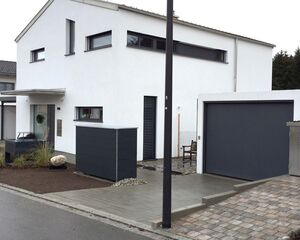 . Modernes weißes Haus mit grauem Garagentor, Vorgarten mit Stein und Pflanzen, gepflasterte Einfahrt.