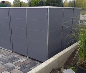 . Ein großes, graues Gehäuse auf einer gepflasterten Fläche, daneben Pflanzen und arbeitende Personen im Hintergrund.