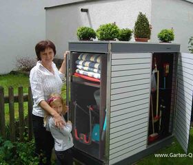 . Frau mit Kind neben Gartenschrank mit Werkzeugen und Tüchern, Pflanzen obendrauf.