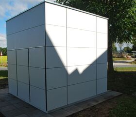 Ein weißer quaderförmiger Bau mit linearem Muster auf einer gepflasterten Fläche, umgeben von Bäumen und Landschaft.