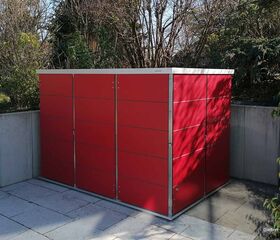 . Ein rotes Gartenhaus steht auf einer gepflasterten Terrasse, umgeben von Bäumen und Sträuchern.