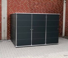 . Ein grauer Metallschuppen steht auf einem gepflasterten Boden vor einer roten Backsteinmauer.