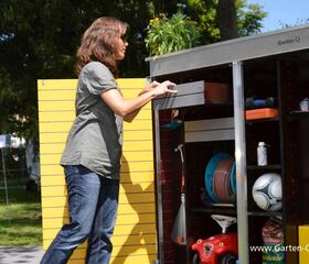 . Eine Frau benutzt einen Gartenschrank mit gelber Tür, darin sind Gartengeräte, ein Ball und Spielzeug.
