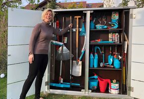 Eine Frau steht neben einem geöffneten Gartenschrank mit verschiedenen Gartengeräten und Werkzeugen.