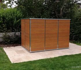 . Ein modernes Holzlagerhaus steht auf einer Betonfläche im Garten, umgeben von Bäumen und Sträuchern.