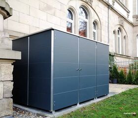 . Ein moderner, grauer Gartenschuppen neben einem Gebäude aus Stein mit großen Fenstern und umgeben von Rasenfläche.