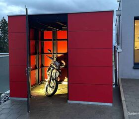 Ein roter Schuppen mit offener Tür beherbergt ein Motorrad. Die Beleuchtung hebt das Fahrzeug hervor.