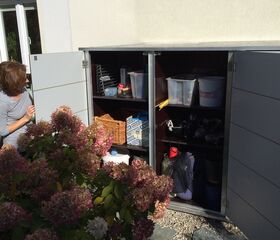 . Ein offener Gartenschrank mit Gartenzubehör und Behältern, daneben eine Frau und blühende Hortensien.