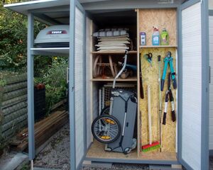 Ein geöffneter Gartenschrank mit Werkzeugen, Reinigungsmitteln, einem Handwagen und einem Dachbox-Kofferraum.