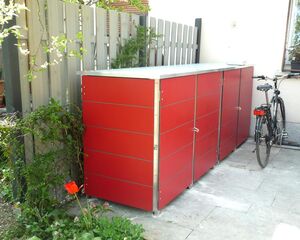 . Ein roter Mülltonnenunterstand neben einem Zaun mit einem Fahrrad und Pflanzen im Hintergrund.