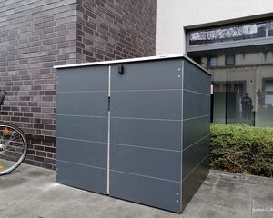 Ein grauer Metallschuppen neben einem Gebäude mit einem Fahrrad im Vordergrund.