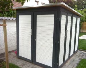 Ein Gartenhaus mit weißen Türen und dunklem Rahmen auf einem gepflasterten Bereich im Freien.