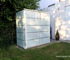 . Ein moderner Gartenschrank aus Glas oder Kunststoff steht neben einem Gebäude und einer Hecke im Freien.