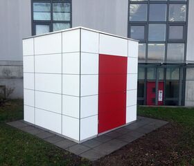 Ein kubisches Gebäude mit weißen Fliesen und einer roten Tür vor einem größeren Gebäude.