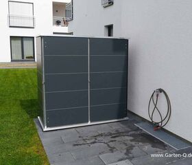 . Eine graue Gartengerätebox steht auf einer Terrasse neben einer Hauswand, mit einem Wasserschlauch an der Wand.