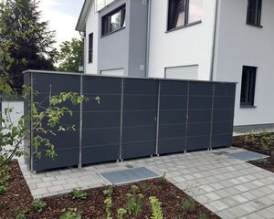 . Ein Zaun um Mülltonnen vor einem modernen Haus auf gepflasterter Fläche mit Garten.
