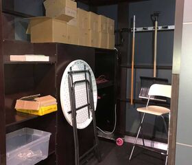 Regale mit Kartons, ein zusammenklappbarer Tisch, Stühle und Gartengeräte in einem Lagerraum.