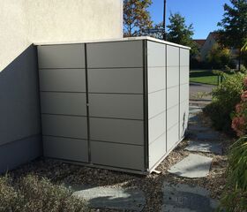 . Ein graues Metall-Gartenhaus an der Hauswand, umgeben von Kies und einem Gartenweg.