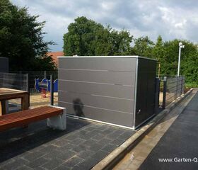 . Ein grauer Schuppen im Freien mit Holzbänken und einem Spielplatz im Hintergrund.