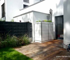 . Moderner Garten mit Holzterrasse, weißem Schrank, schwarzen Zäunen und Pflanzen.