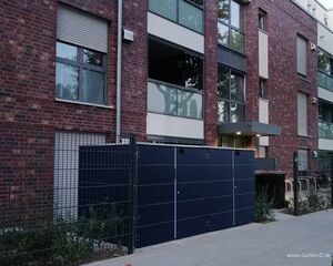 Ein mehrstöckiges Gebäude mit Backsteinfassade und großen Fenstern, daneben Metallzäune und ein Weg.