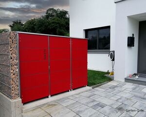 . Rotes Paketlager vor einem Haus mit Steinmauer, großer Sichtschutz neben Einfahrt.