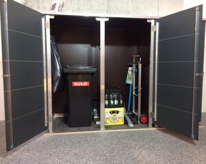 . Ein geöffneter Schrank mit einem Müllcontainer, einer Kiste mit Flaschen und einem Handwagen.