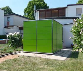 . Ein grüner Würfel im Garten, umgeben von Rasen, Blumen und einem Gebäude im Hintergrund.