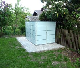 . Ein moderner, rechteckiger Schuppen mit Glaswänden steht auf einer gepflasterten Fläche im Garten.