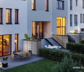 . Moderne Terrasse mit Tisch und Stühlen, umgeben von Hecken, vor einem beleuchteten Gebäude.